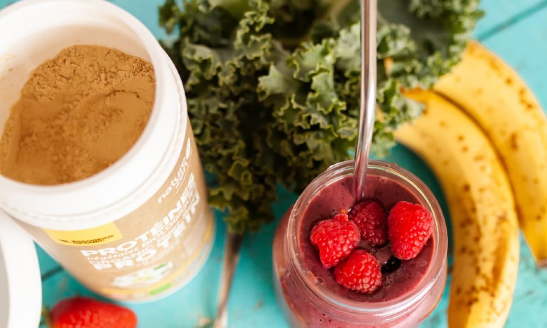 A canister of protein drink powder next to a smoothie glass with berries.